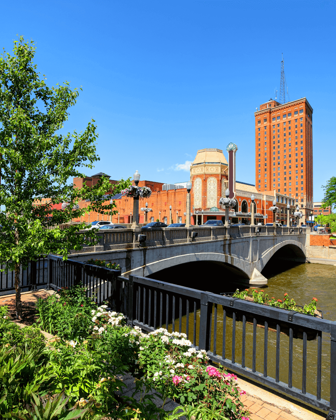 Scenic downtown view with historic bridge over river, red-brick buildings, and colorful flower beds in the foreground