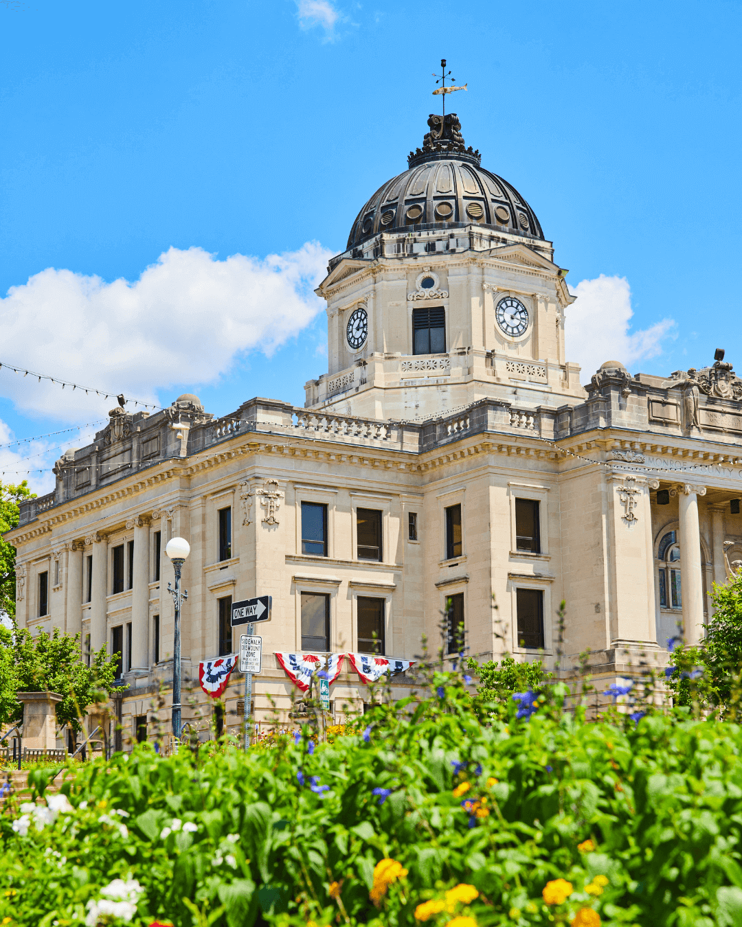 Historic courthouse-style building with clock tower and patriotic decorations, surrounded by greenery and flowers